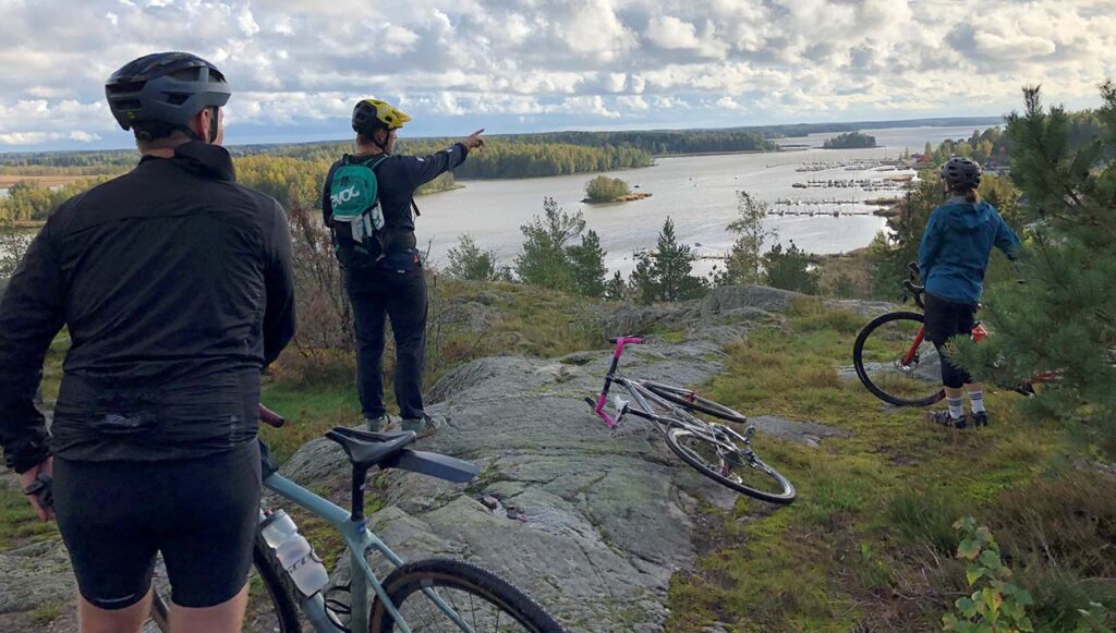 [Alt-teksti: kolme pyöräilijää on pysähtynyt kumpuilevan matalan kallion päälle katsomaan maisemia järven suuntaan.]