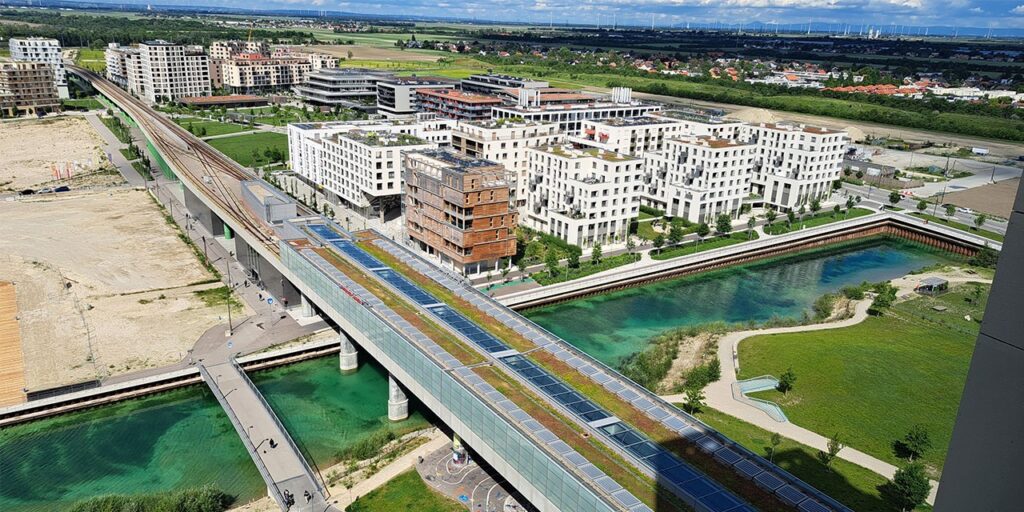 A photo from outdoor with a metro line crossing a new city area. In front, there is a manmade lake, behind the lake, several modern buildings stand.