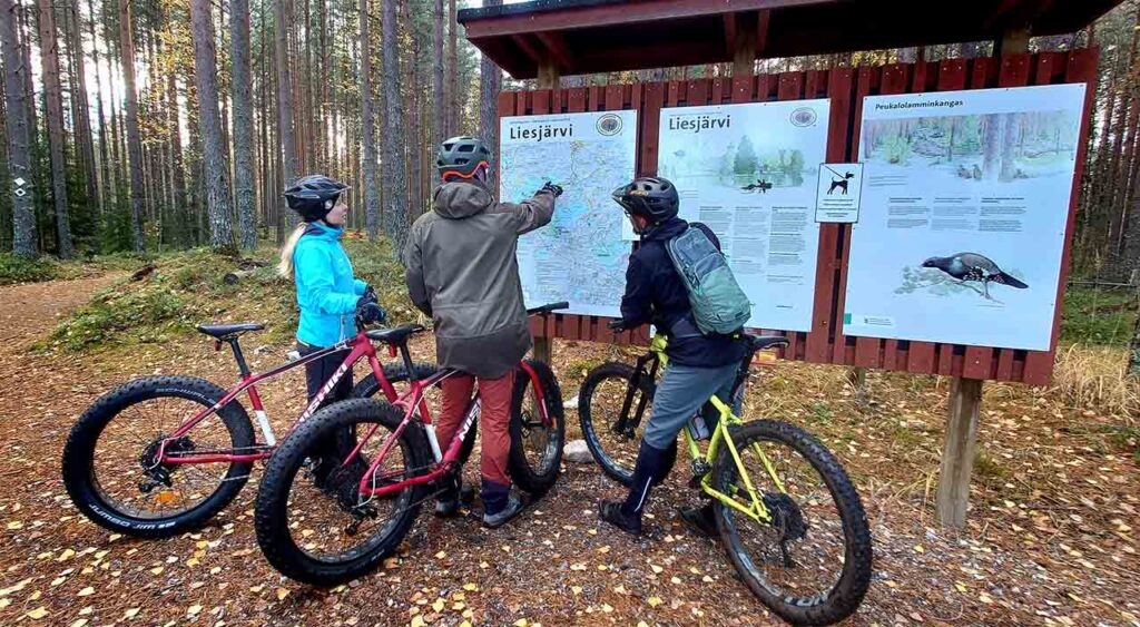 [Alt-teksti: kolme henkilöä maastopyörien vierellä tutkimassa opastetaulua metsässä.]