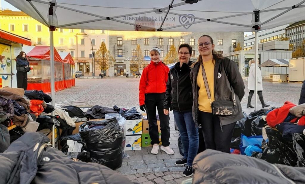 [Alt-teksti: Lahden torille pystytetty tapahtumakoju, jolla hymyilee kolme naista, heidän ympärillään on säkeissä ja laatikoissa paljon vaatetavaraa.]