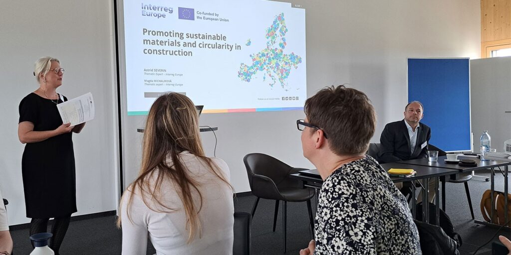 An indoor photo of people sitting and listening to a female speaker. In the centre, there is a screen with a title of Promoting sustainable materials and circularity in construction, Interreg Europe logo and a map of Europe.