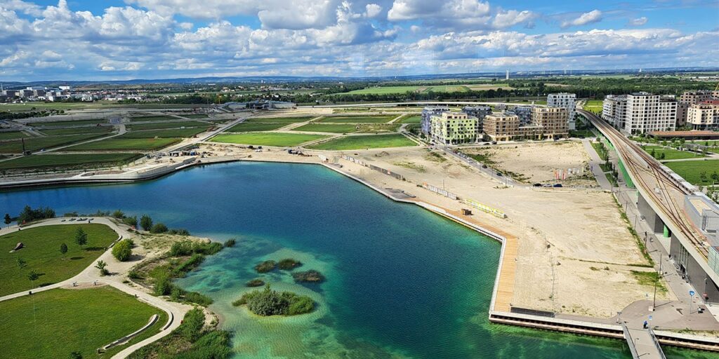 An aerial view on a district, which is under construction and built around a man-made lake. on the right, there is the metro/train line. It is a sunny day with white clouds on the sky.