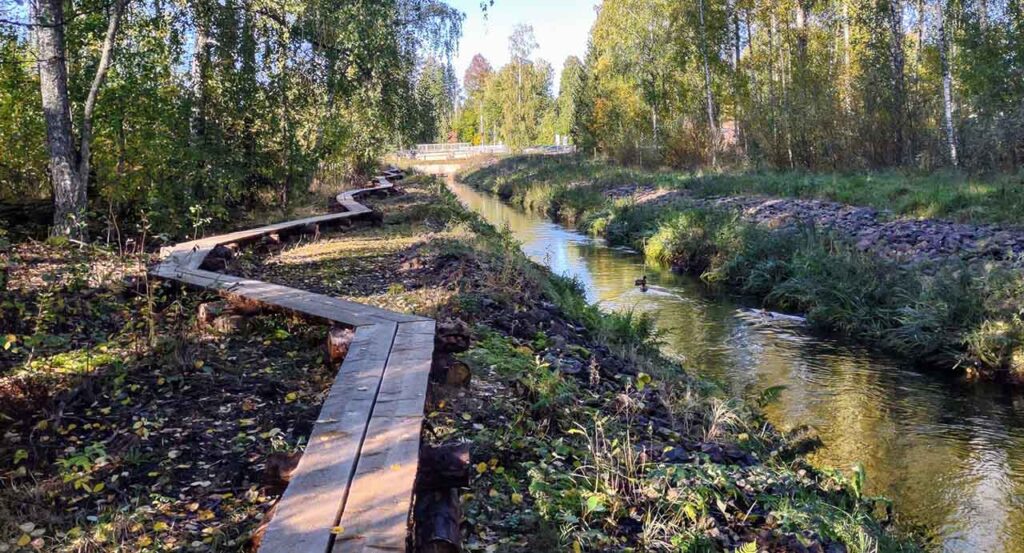 [Alt-teksti: syksyinen maisema lehtipuineen, oja, jossa virtaa vettä, ojan vieressä rannalla kulkee hieman mutkitteleva pitkospuupolku.]