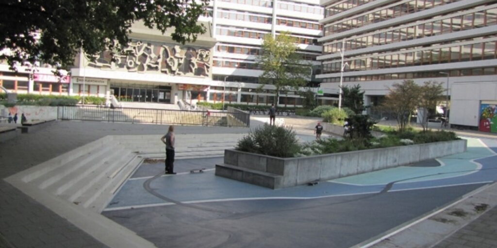 An open concrete area with steps going up to street level, there are 3 people standing within it and a raised central area containing plants.