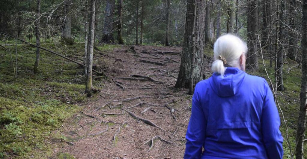 [Alt-teksti: metsäinen polku, jota pitkin kävelee naisoletettu henkilö.]