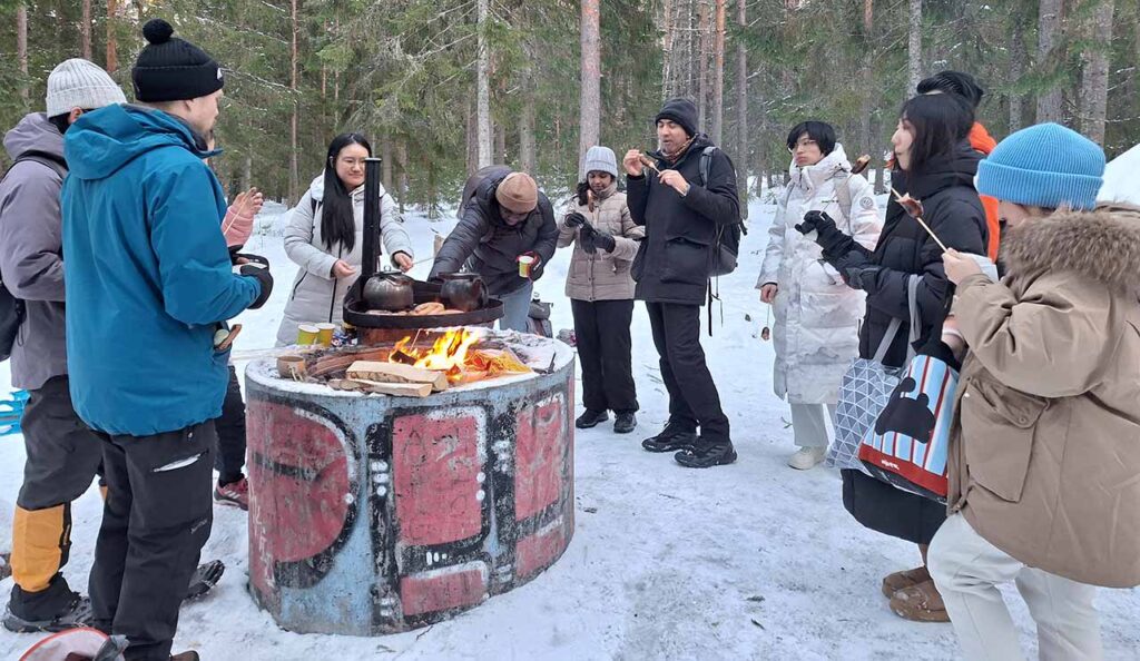 [Alt-teksti: Joukko ihmisiä on nuotion ympärillä  ruokailemassa metsässä talvella.]