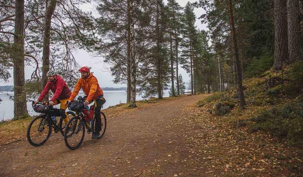 [Alt-teksti: Kaksi henkilöä pyöräilee metsätiellä, joka on vesistön äärellä.]