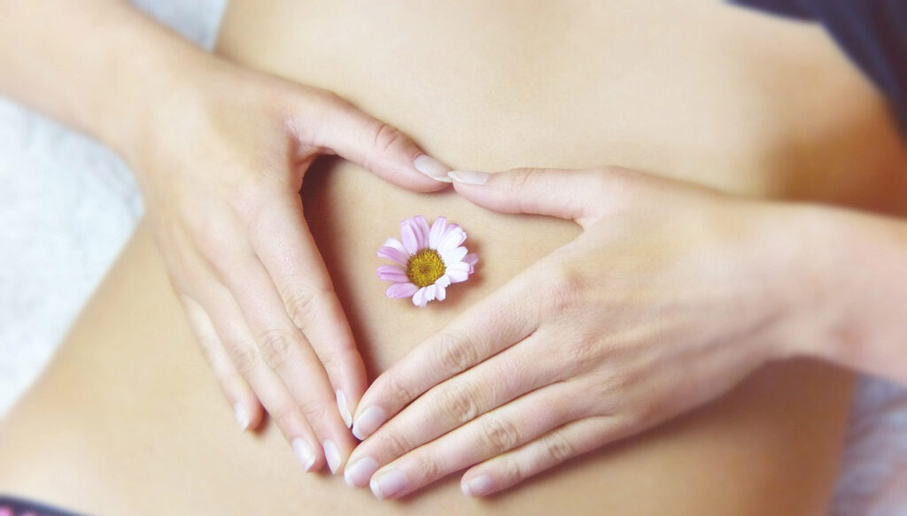[Alt-text:  Woman holding a flower on her stomach, with her hands forming a heart shape around it.]