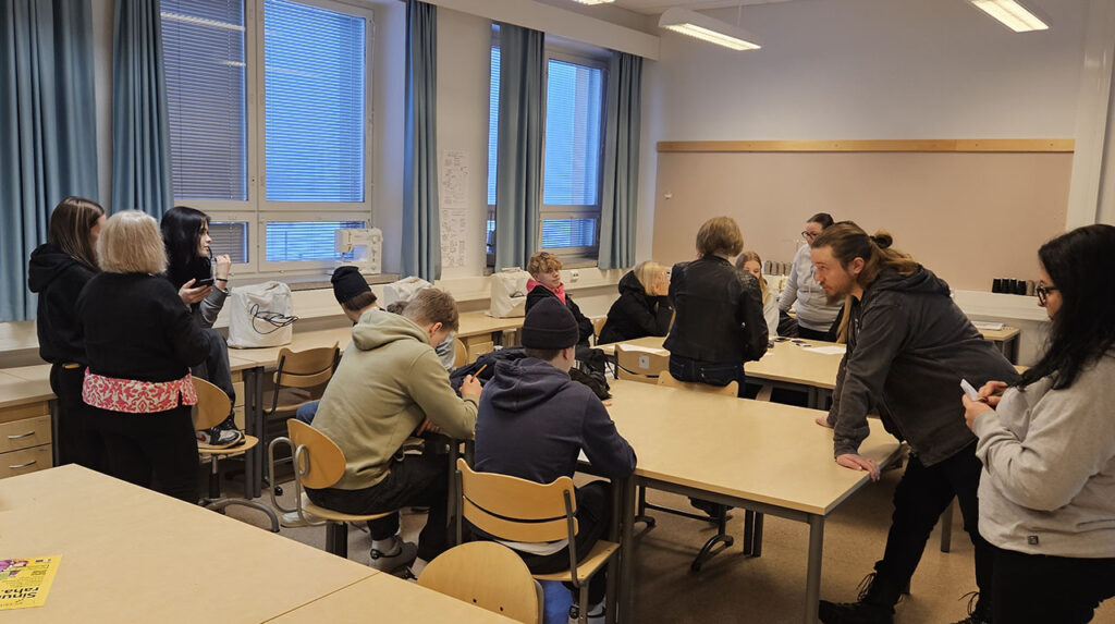 [Alt-teksti: luokkahuone, jossa istumassa ja keskustelemassa nuoria henkilöitä.]