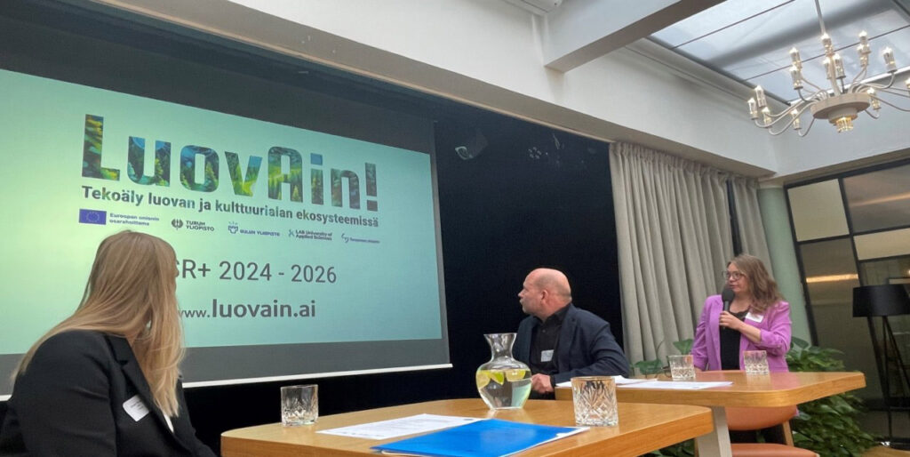[Alt-teksti: henkilö pitämässä presentaatiota, näyttöseinällä teksti luovain tekoäly luovan ja kulttuurialan ekosysteemissä.]
