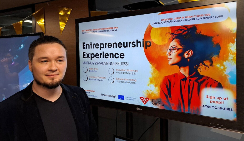 [Alt-teksti: hymyilevä henkilö suuren näyttöruudun edessä, ruudulla lukee entrepreneurship experience sekä muuta tekstiä.]