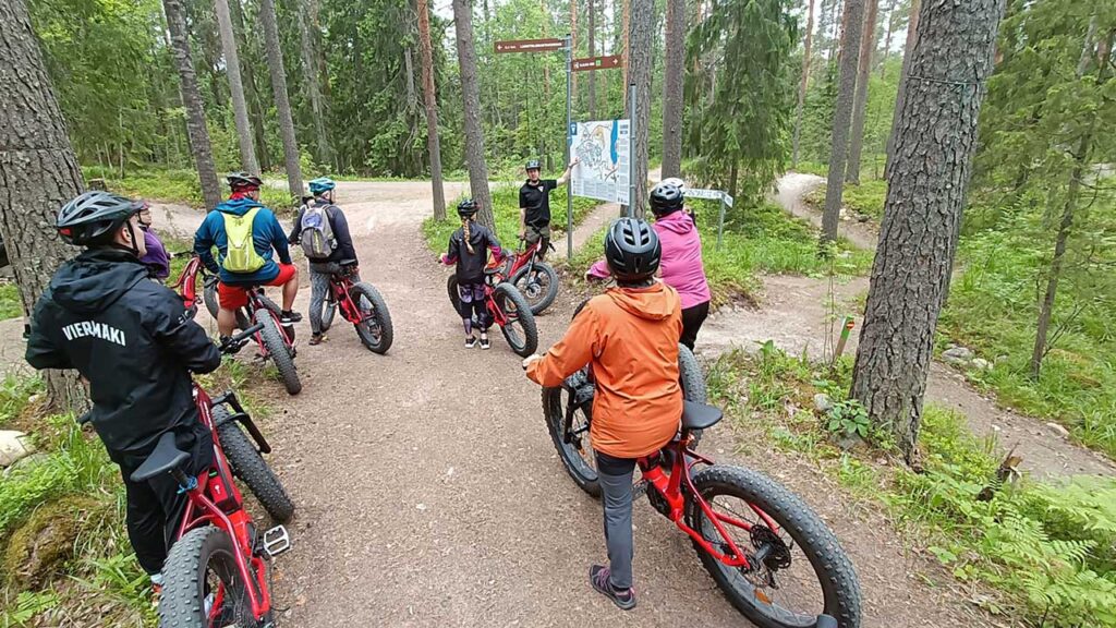 [Alt-teksti: ryhmä pyöräilijöitä kuuntelemassa oppaan selitystä opastustaulun lähellä metsätiellä, ympärillä puunrunkoja ja metsäpolkuja.]