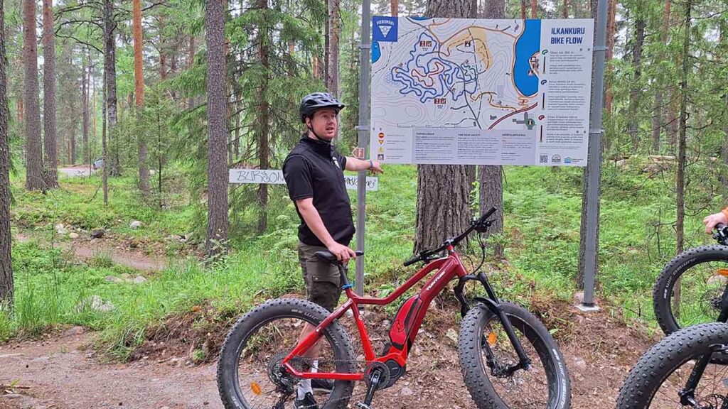 [Alt-teksti: opashenkilö pyöräilykypärä päässä ja maastopyörä vierellään kertoo opastintaulun äärellä metsässä.]