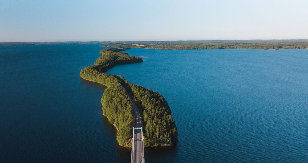 [Alt-teksti: kesäistä järvenselkää halkova metsäinen kannas ja maantie.]