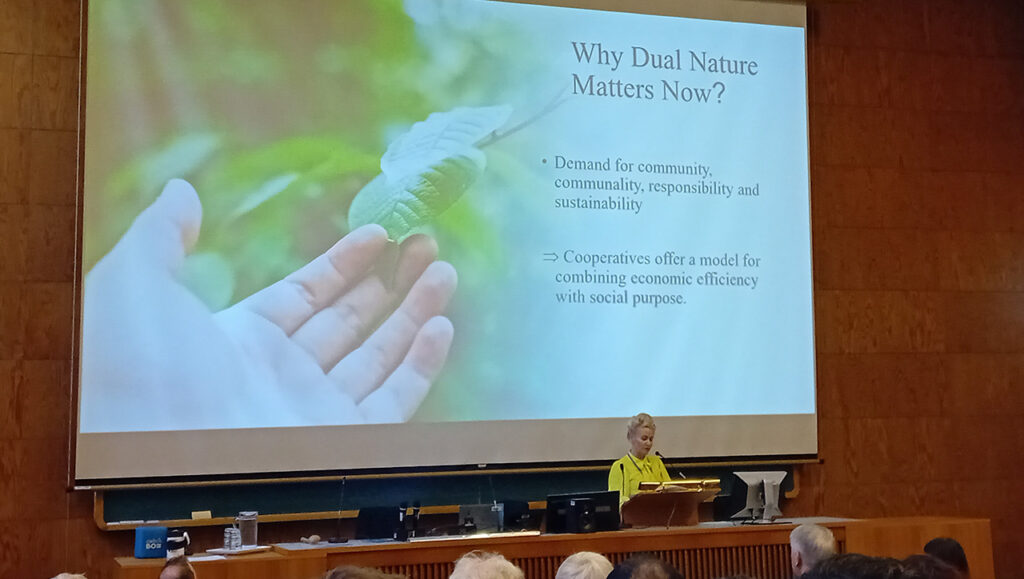 [Alt-teksti: henkilö pitää presentaatiota konferenssisalissa, taustalla näkyy esityksen otsikko why dual nature matters now.]
