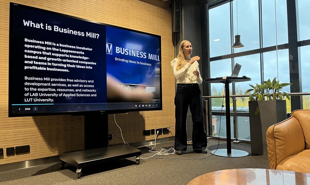 [Alt-teksti: henkilö pitämässä presentaatiota, vieressä suuri näyttötaulu, jossa lukee what is business mill.]