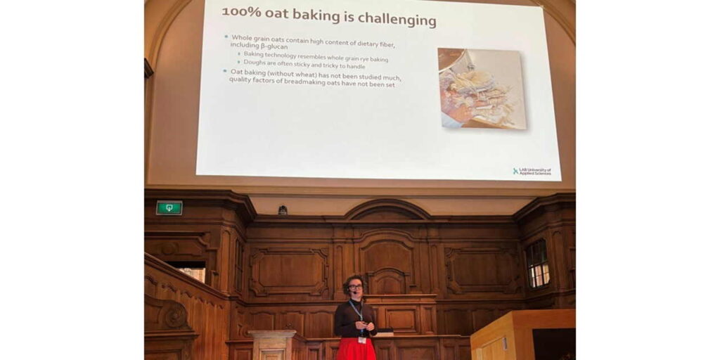 Henkilö pitämässä konferenssiesitystä. Taustalla dia, jossa lukee "100% oat baking is challenging".