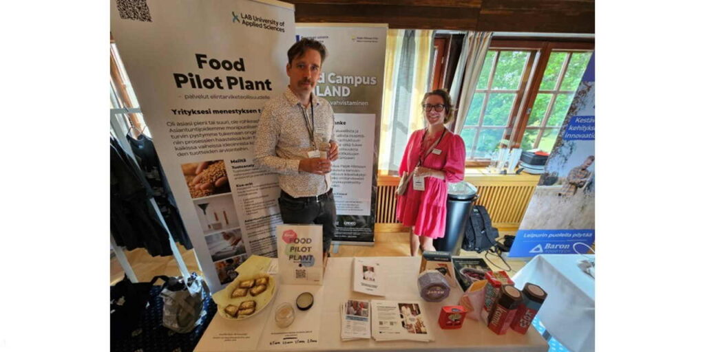 Kaksi henkilöä seisoo pöydän takana. Pöydällä on esitteitä ja erilaisia elintarvikepakkauksia.