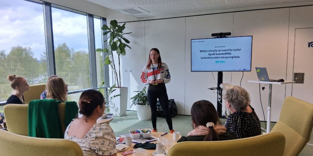 Nainen seisoo pitämässä esitystä valoisassa kokoushuoneessa, jossa on suuret ikkunat. Hän on kasvotusten pöydän ympärillä istuvan yleisön kanssa. Näytöllä näkyy esitys.