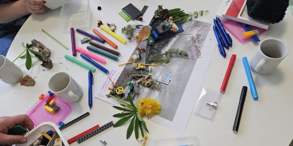 Pöydällä on levällään erivärisiä kyniä, LEGO-palikoita, kasveja ja muita luonnonmateriaaleja. Keskellä pöytää on mustavalkoinen A3-kokoinen valokuva kaupunkimaisemasta, jonka päälle on aseteltu LEGO-palikoita ja luonnonmateriaaleja sekä kirjoitettu muistiinpanoja käsin.