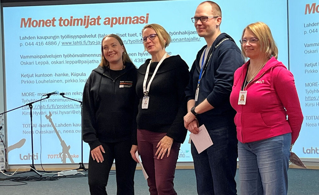 [Alt-teksti: neljä henkilöä seisomassa vierekkäin, heidän takanaan näyttötaululla suuri teksti monet toimijat apunasi.]