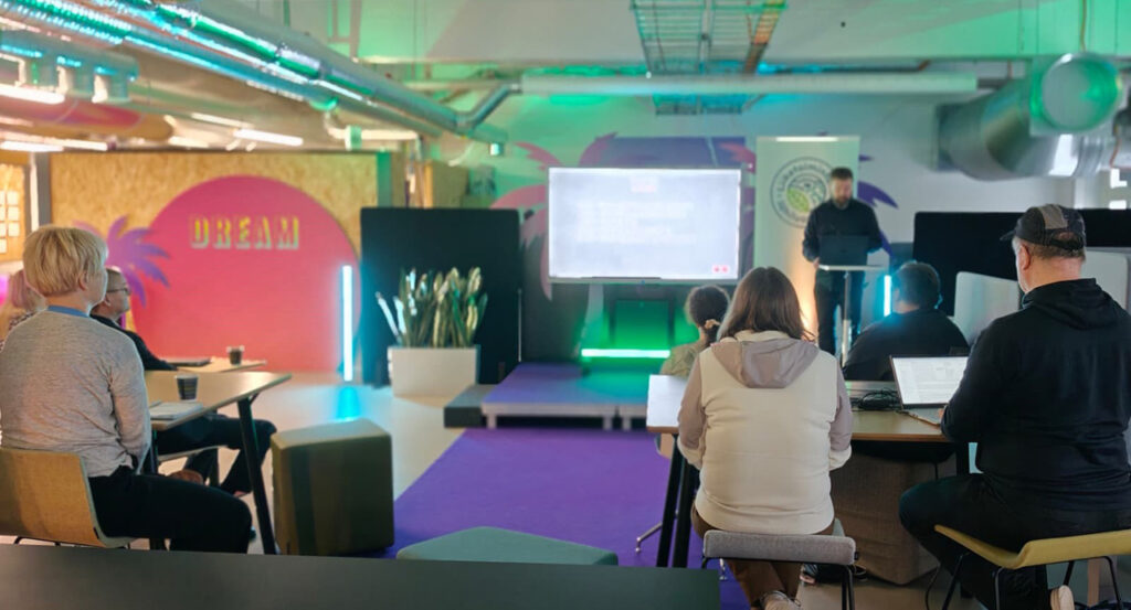 [Alt-teksti: ihmisiä värikkäässä luentotilassa, jossa on pöytiä ja tuoleja.]