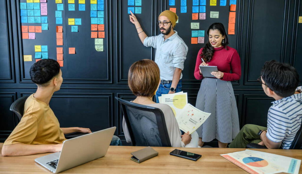 Two people are standing in front of a dark wall that is full of Post-It notes. Three people are looking at them and listening.