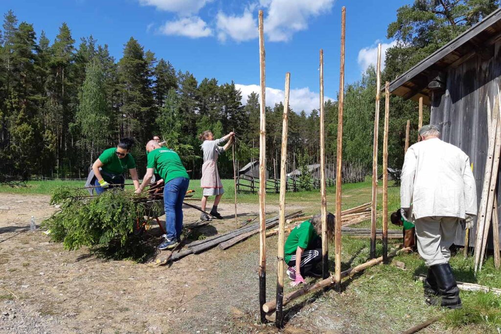 [Alt-teksti: kesäinen maalaispihapiiri, jossa työskentelee ihmisiä pystyttäen tukirunkoja.]