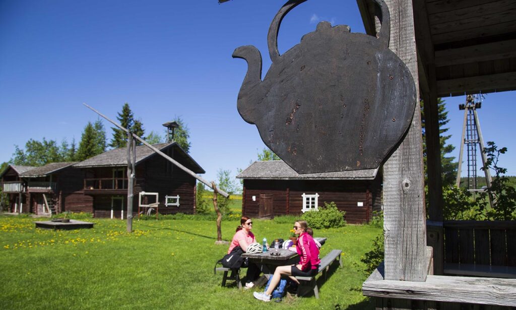 [Alt-teksti: maalaismainen pihapiiri, jossa on useita vanhoja rakennuksia sekä pihalla ihmisiä pitämässä kahvitaukoa.]