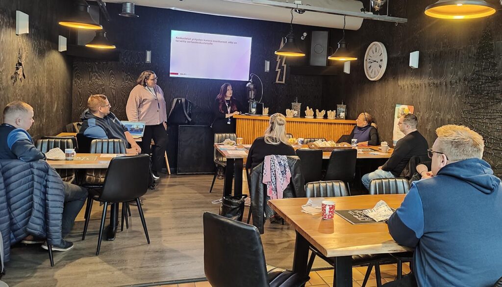 [Alt-teksti: kokoustila, jossa on ihmisiä istumassa pöydissä ja kuuntelemassa kahden henkilön pitämää esitystä.]