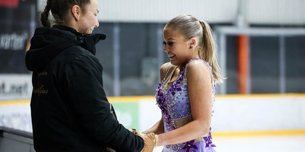 [Alt-teksti: valmentaja mustassa takissa pitää käsistä kiinni kimaltavaan kisa-asuun pukeutunutta taitoluistelijaa.]