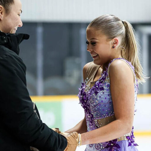 [Alt-teksti: Mustaan urheilutakkiin pukeutunut valmentaja pitää käsistä kiinni kimaltavassa kisa-asussa olevaa taitoluistelijaa.]