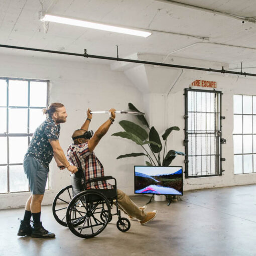 [Alt-teksti: henkilö tukee pyörätuolissa istuvaa henkilöä, samalla kun pyörätuolissa istuva pelaa VR laseilla.]