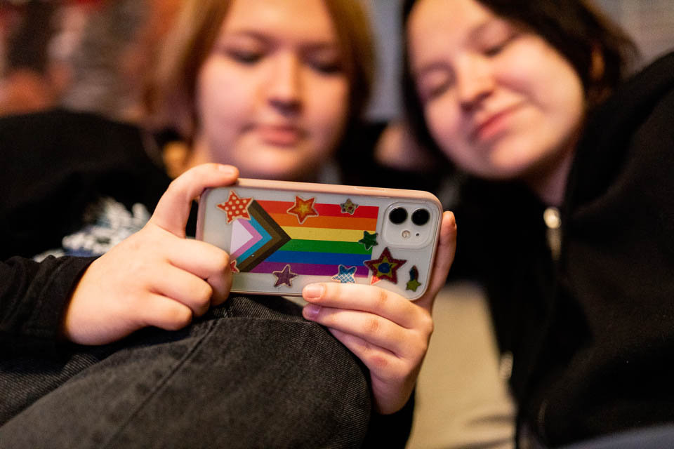 [alt-teksti: kaksi nuorta katsoo jotain puhelimen ruudulta, jossa on sateenkaaritausta.]