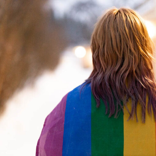 [Alt-teksti: sateenkaarilipun alle kääriytynyt ihminen.]