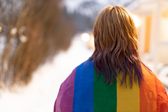 [Alt-teksti: sateenkaarilipun alle kääriytynyt ihminen.]