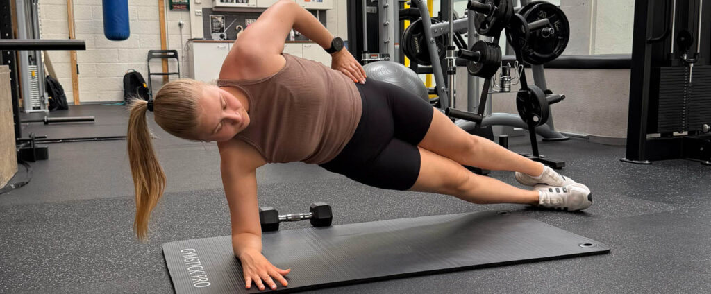 [Alt-teksti: nainen kuntosalin lattialla tekemässä pitkittäissuuntaista harjoitetta, joka kohdistuu keskivartalon ja lonkan alueelle.] 