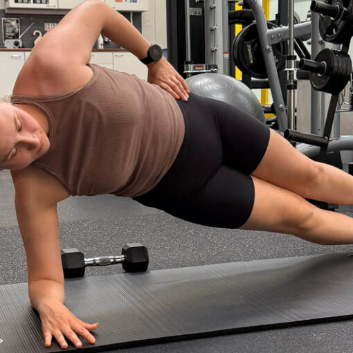 [Alt-teksti: nainen kuntosalin lattialla tekemässä pitkittäissuuntaista harjoitetta, joka kohdistuu keskivartalon ja lonkan alueelle.]