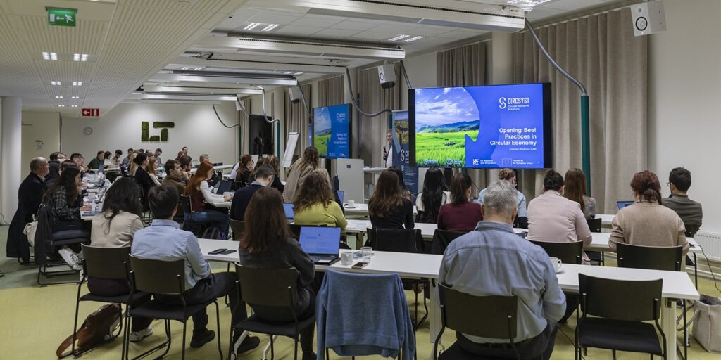 Ihmisiä istumassa pöytien ääressä ja kuuntelemassa puhujaa. Taustalla näyttö, jossa lukee CircSyst.