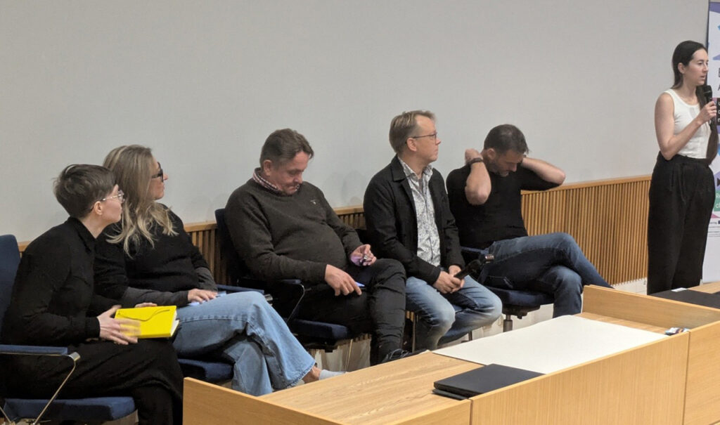 [Alt-teksti: Viisi panelistia istuu tuoleilla ja paneelin vetäjä esittää kysymystä.]