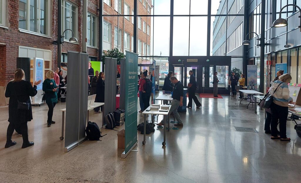 [Alt-teksti: korkea lasiseinäinen aulatila, jossa on ihmisiä liikkeellä tutkimassa esittelypöytiä ja seisovia julisteita.]