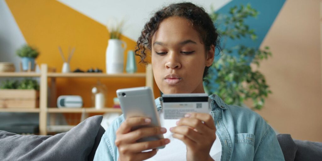 Afroamerikkalainen tyttö hymyilee maksaessaan verkossa pankkikortilla ja älypuhelimella kotona, nauttien ostoksista kotoa käsin.