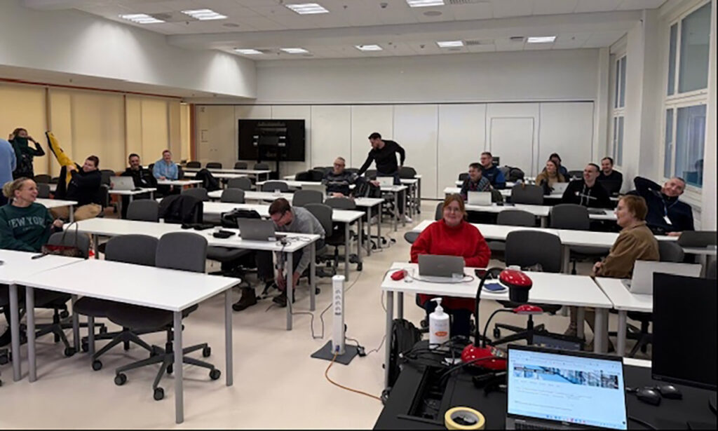 [Alt-teksti: luokkatila, johon on asettumassa ihmisiä pöytien ääreen kuuntelemaan.]