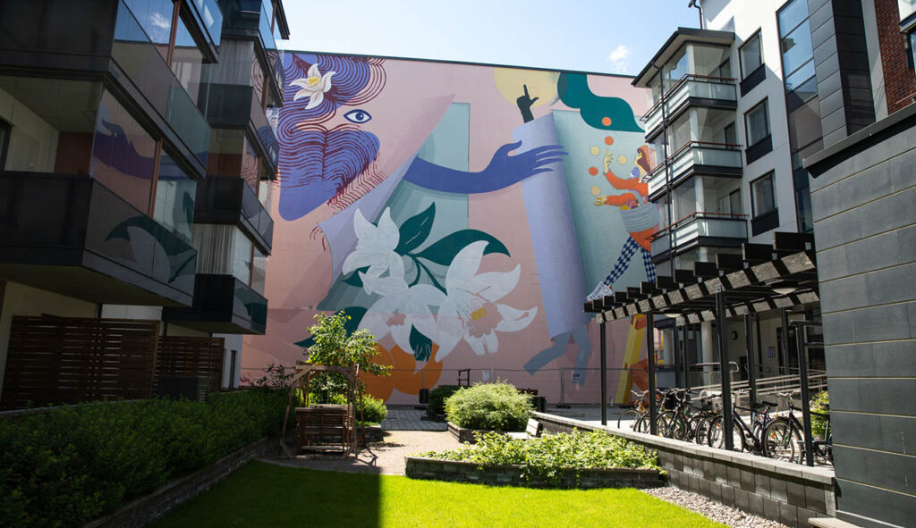 [Alt-teksti: kerrostalojen välinen sisäpiha, takana korkean rakennuksen seinä, jolle on maalattu monivärinen taideteos.]