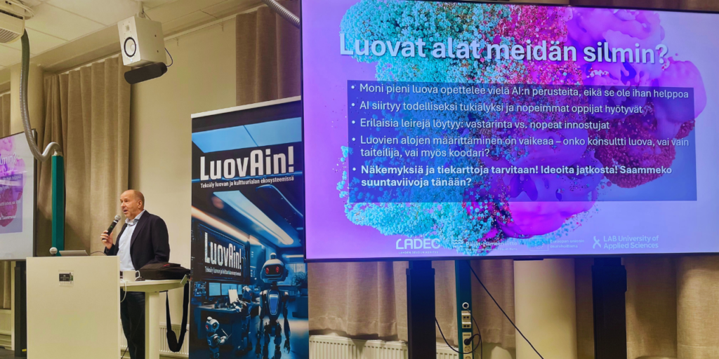 Esiintymistilanne tapahtumassa, jossa käsitellään luovien alojen ja tekoälyn roolia. esiintyjä puhumassa ja etualalla näkyy suuri näyttö, jossa otsikko 'Luovat alat meidän silmin?' ja listattuja teemoja.