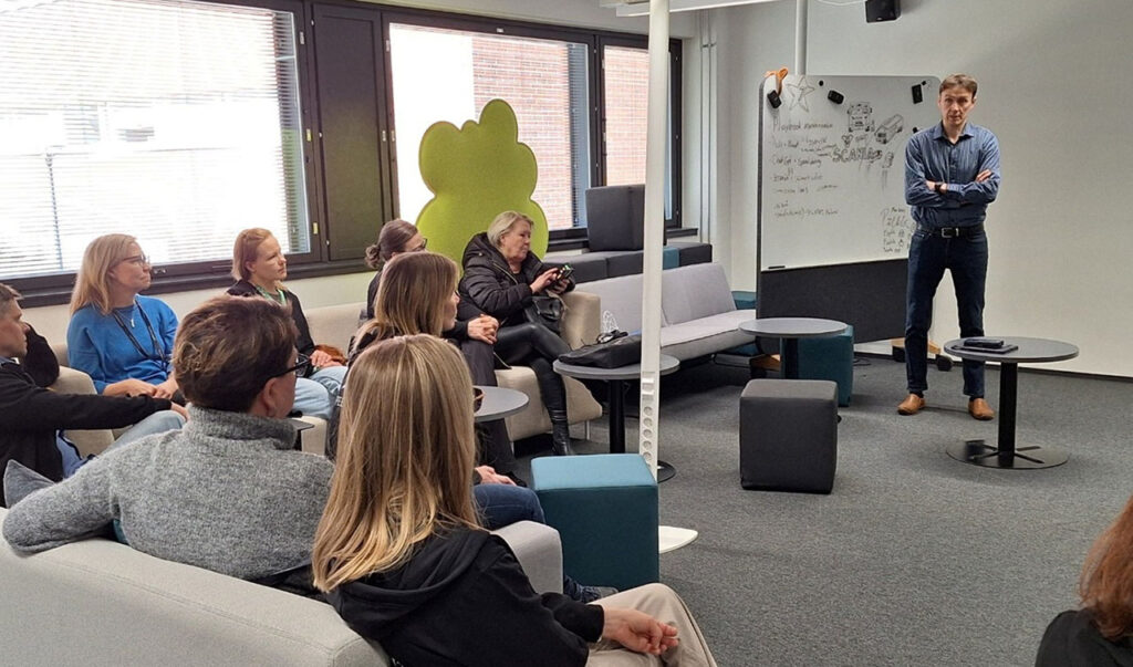 [Alt-teksti: suuri ryhmätyöskentelytila, jossa istuu ihmisiä kuuntelemassa yhden henkilön pitämää esitystä.]