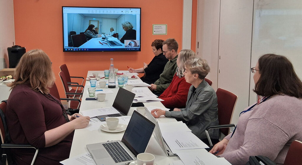 [Alt-teksti: simulaatiotilanne seminaarihuoneessa, pitkän pöydän ääressä istuu henkilöitä, seinällä on skriini, jossa näkyy myös työskenteleviä henkilöitä.]