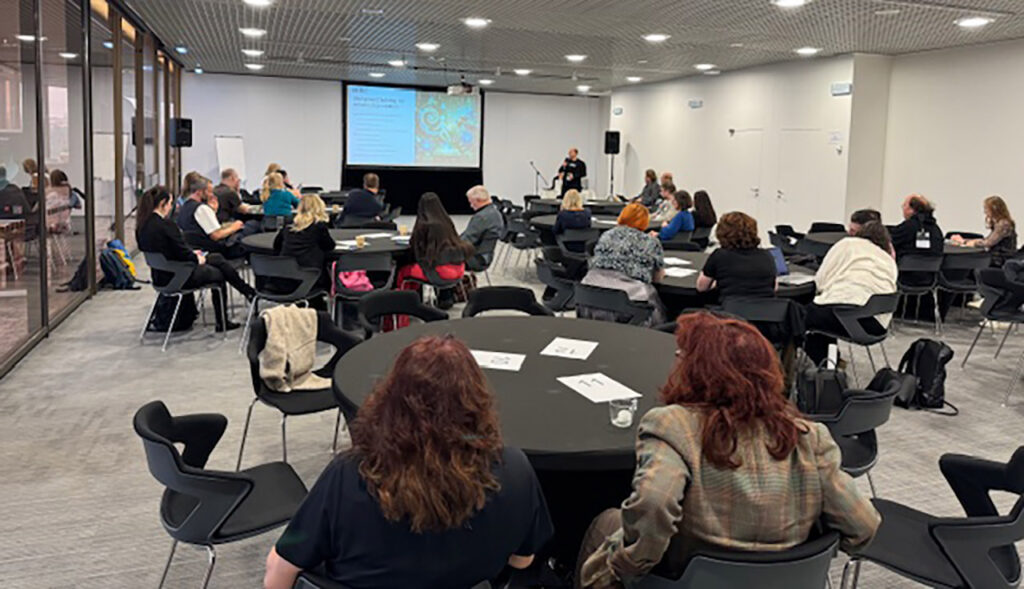 [Alt-teksti: suuri opintosali, jossa on pyöreiden pöytien ääressä ihmisiä työskentelemässä, salin etuosassa henkilö pitää esitystä.]