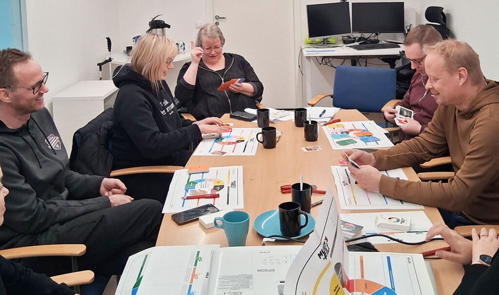 [Alt-teksti: hymyileviä henkilöitä istumassa pitkän pöydän ääressä, pöydällä on värikkäitä papereita, kahvimukeja, kyniä ja muuta tavaraa.]