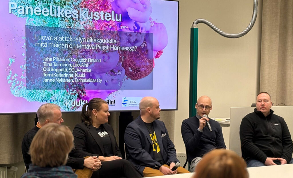 [Alt-teksti: viisi henkilöä istumassa vierekkäin pöydän ääressä, yksi henkilöistä puhuu mikrofoniin.]